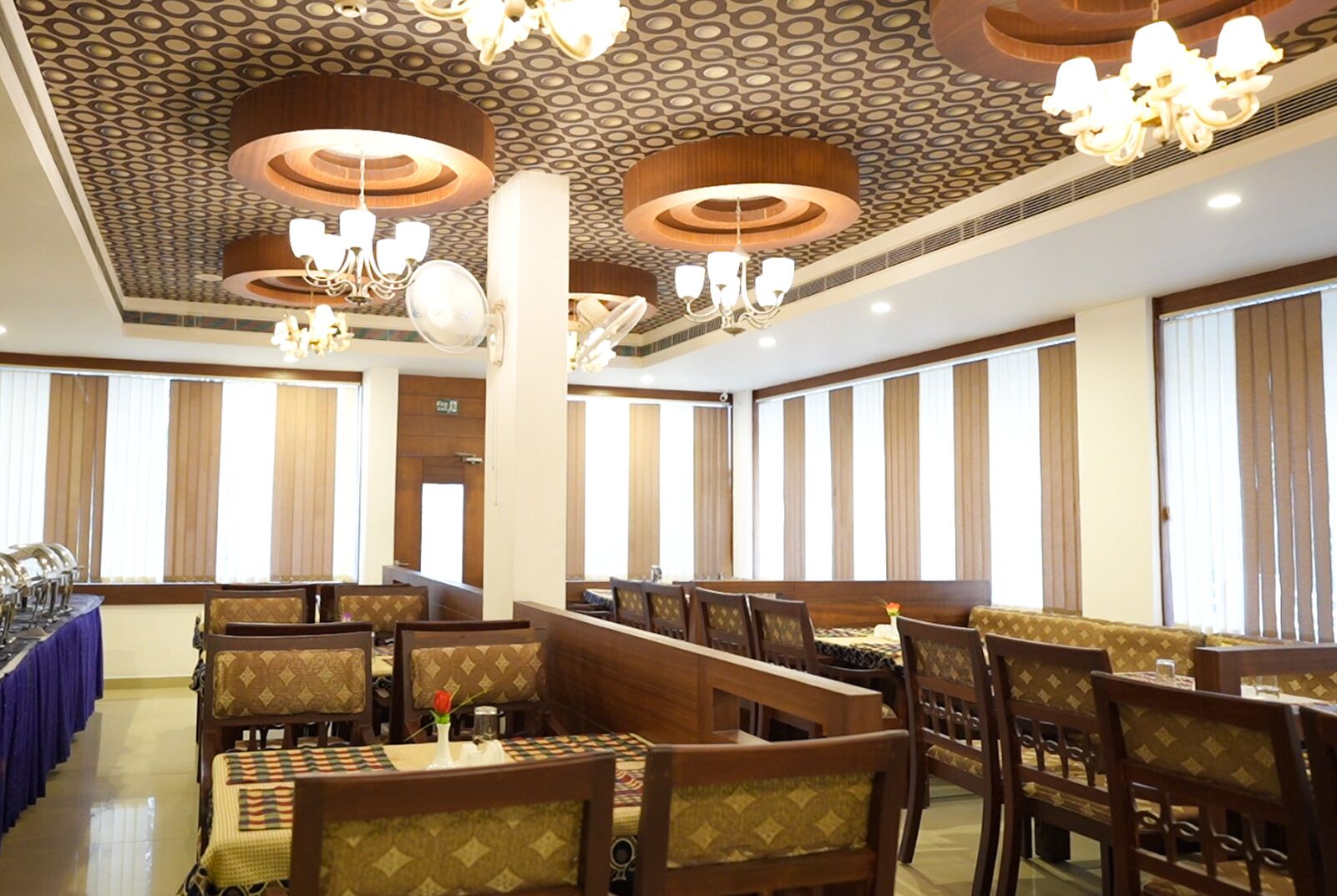 Comedor con varias mesas y sillas, techo estampado y ventanas grandes con cortinas.