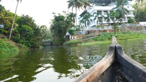 Vista de un río con un bote de madera en primer plano, un edificio blanco con balcones y palmeras al fondo.