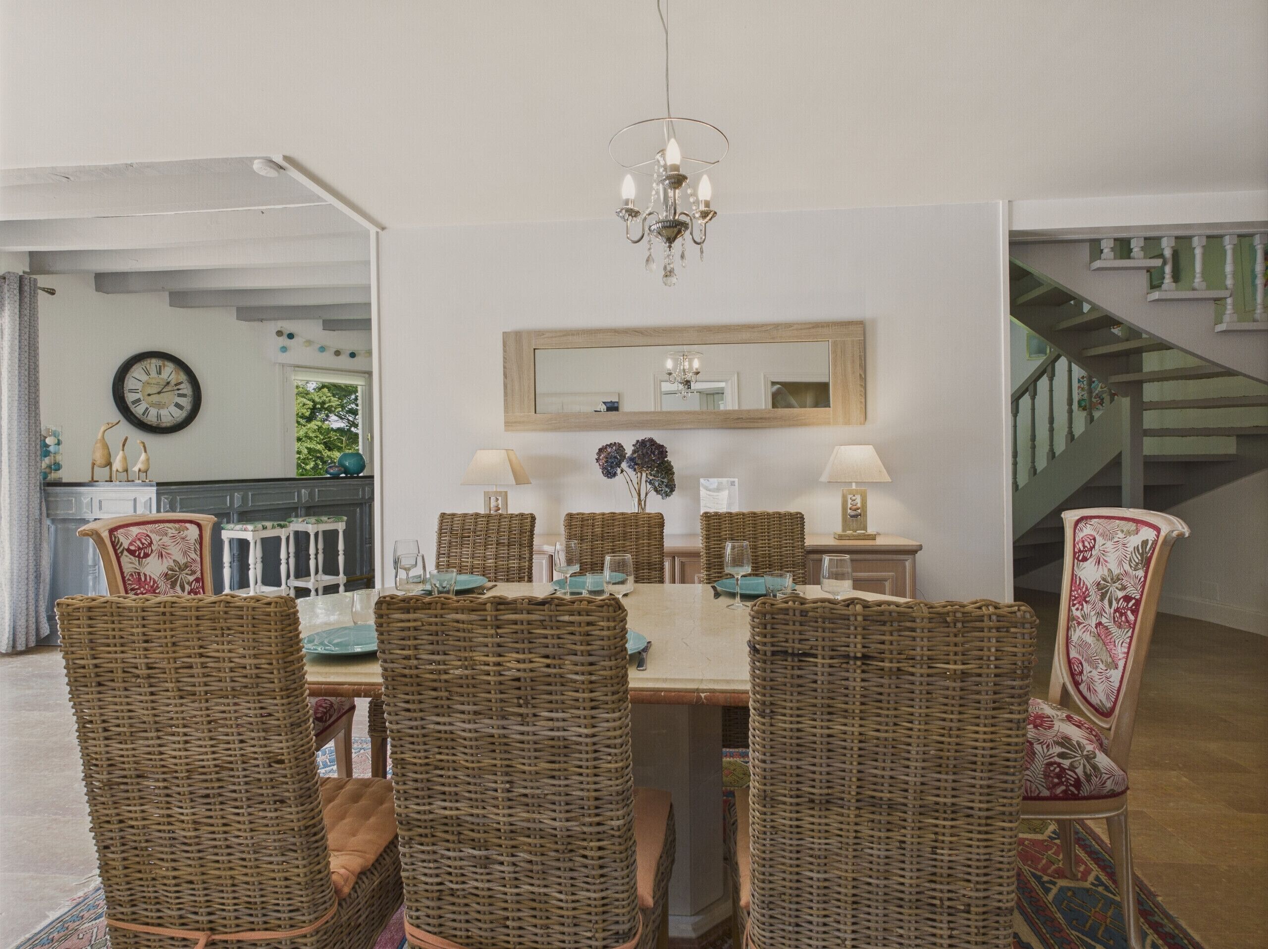 Een eetruimte met een tafel voor zes personen, omringd door rieten stoelen, een kroonluchter en een spiegel boven de tafel.