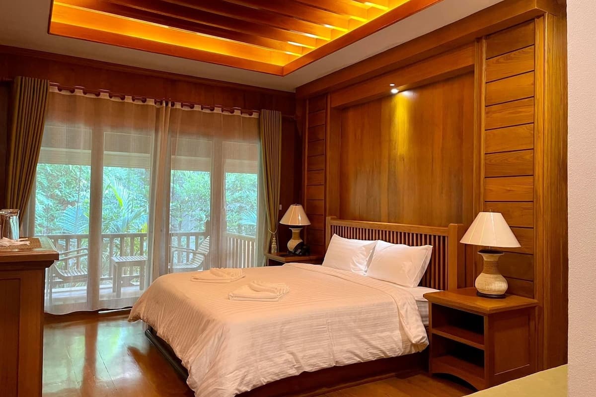 A hotel room with a wooden ceiling, a bed with white linens, two bedside lamps, and a view of greenery through the window.