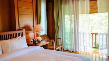 A hotel room with a large bed, a bedside table with a lamp, a chair, and a view of greenery through sheer curtains.