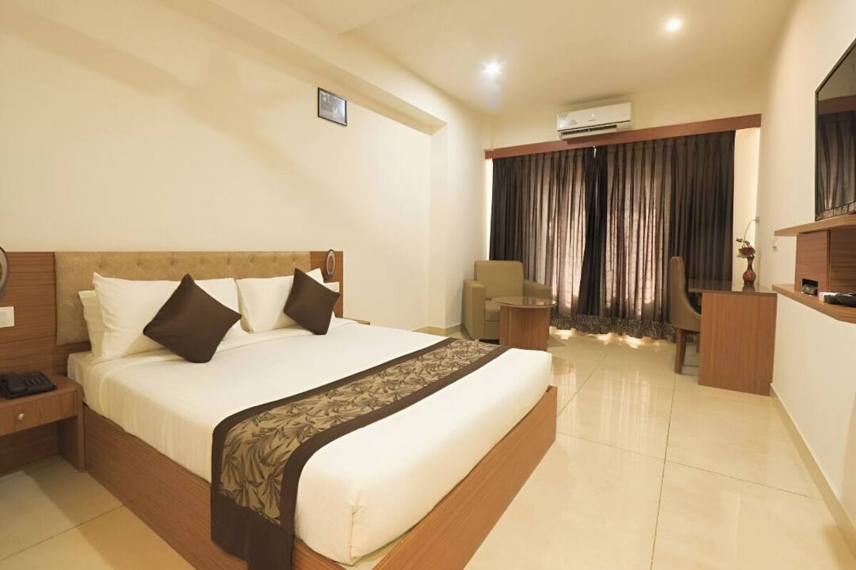 A hotel room with a large bed, bedside tables, a desk, a chair, and an air conditioning unit.