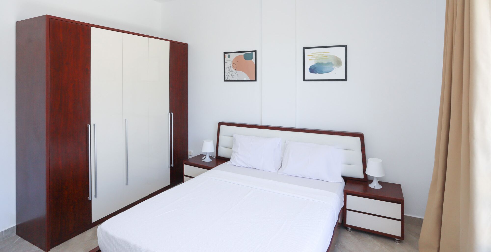 A hotel room with a bed, nightstands, a wardrobe, and two framed artworks on the wall.