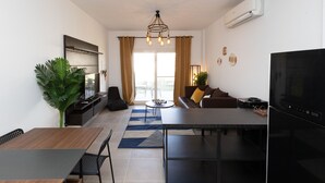 A modern living room with a sofa, coffee table, and a TV. There is a dining area with a table and chairs, and a kitchen area with a refrigerator and microwave. - Prime Holidays Amwaj (El Alamein)