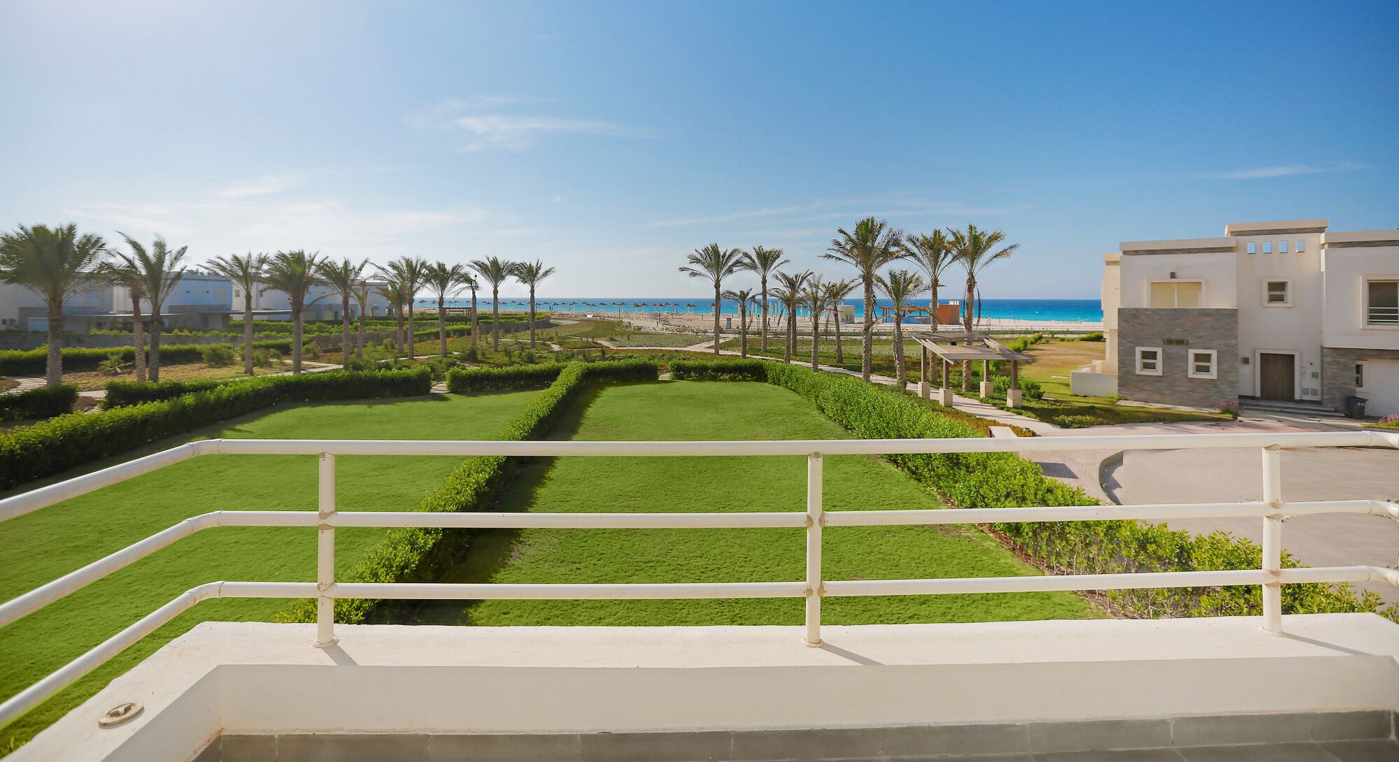 Eine angelegte Grünfläche mit einem Weg, Palmen und Blick auf Strand und Meer.