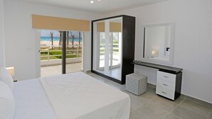 A modern bedroom with a bed, a bedside table, a mirror, and a view of the beach through a sliding glass door. - Prime Holidays Amwaj (El Alamein)