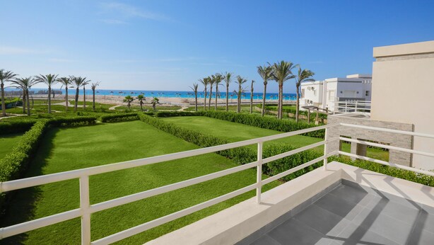 A balcony with a view of a well-maintained lawn, palm trees, and a beach with umbrellas. - Prime Holidays Amwaj (El Alamein)