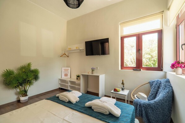 Habitación de hotel con cama, televisor montado en la pared, una mesita con botella y fruta, una silla con una manta y una ventana con vistas a árboles.