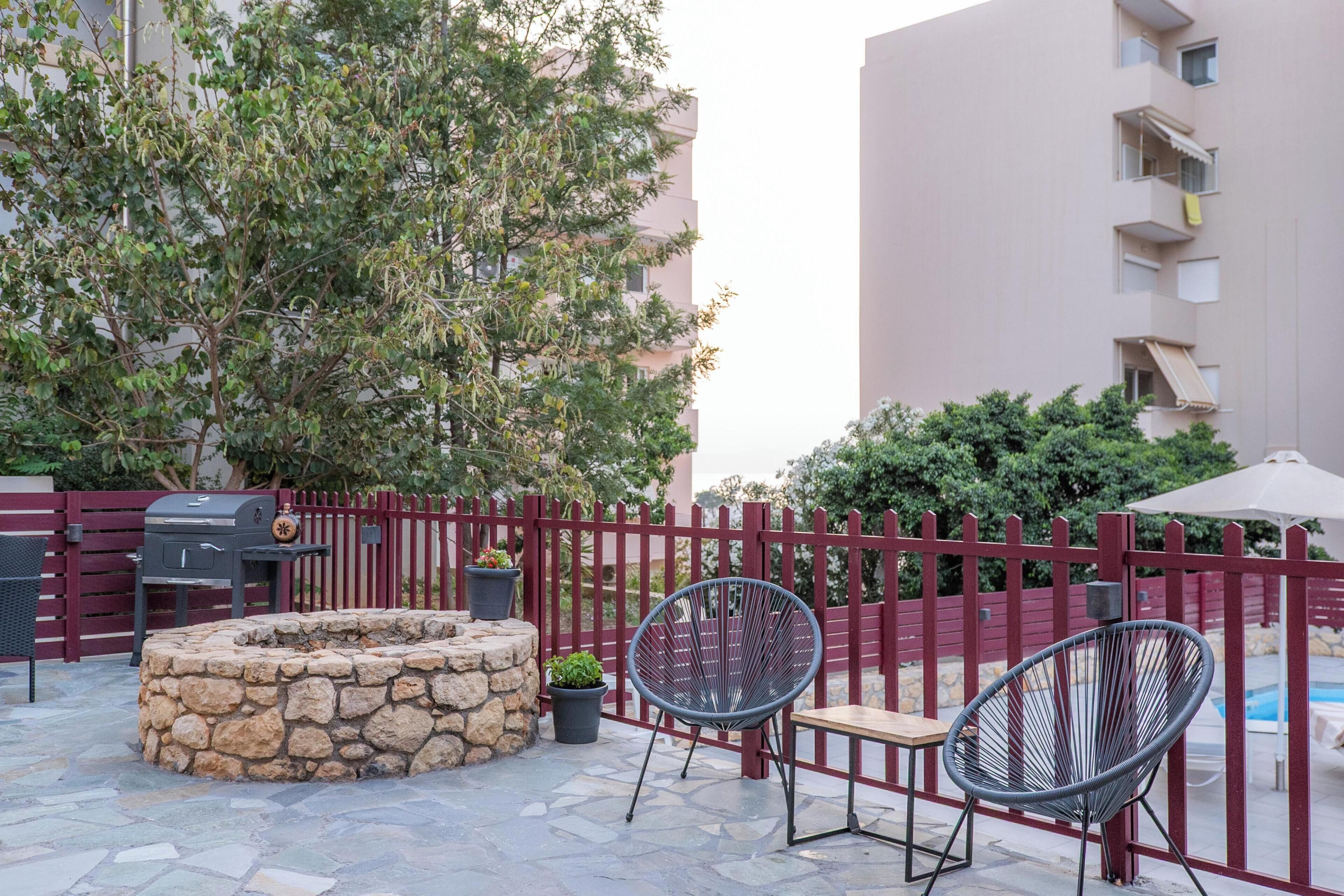 An outdoor patio with stone fire pit, two chairs, a grill, and a pool in the background.