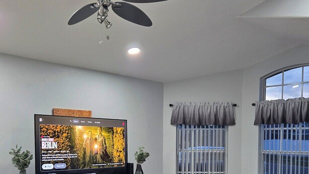 A modern living room with a flat-screen TV, a ceiling fan, a fireplace, and a grey sofa.