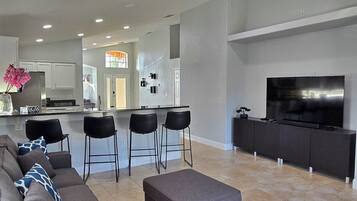 Un salon moderne avec un canapé d’angle gris, un pouf gris et un téléviseur à écran plat posé sur un meuble noir. On y trouve également deux tabourets de bar noirs et un ventilateur de plafond.