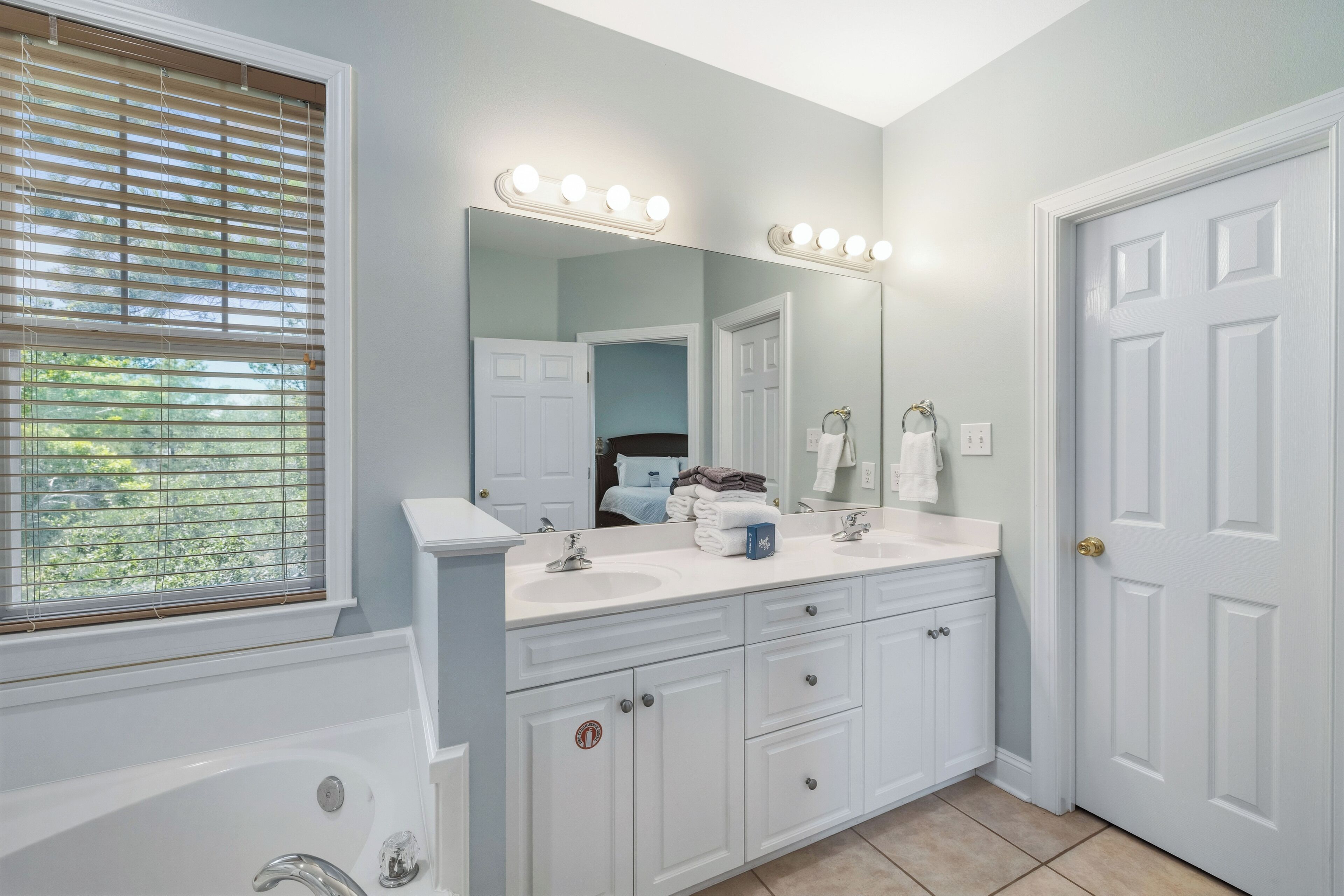 A bathroom with a white vanity, a large mirror with lights, a bathtub, and a door.