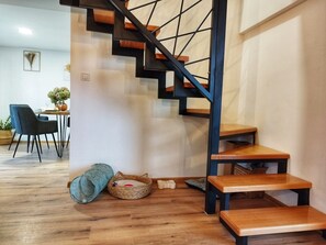 Stairs lead to an upper level, with a dining area below featuring a table for two and comfortable seating.