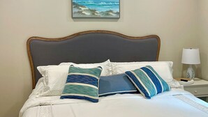 A neatly made bed with white linens and striped pillows, a framed painting of a beach scene above it, and a bedside table with a lamp and a vase. - Palm Cottage, at Magnolia Court Suites (Beaufort)