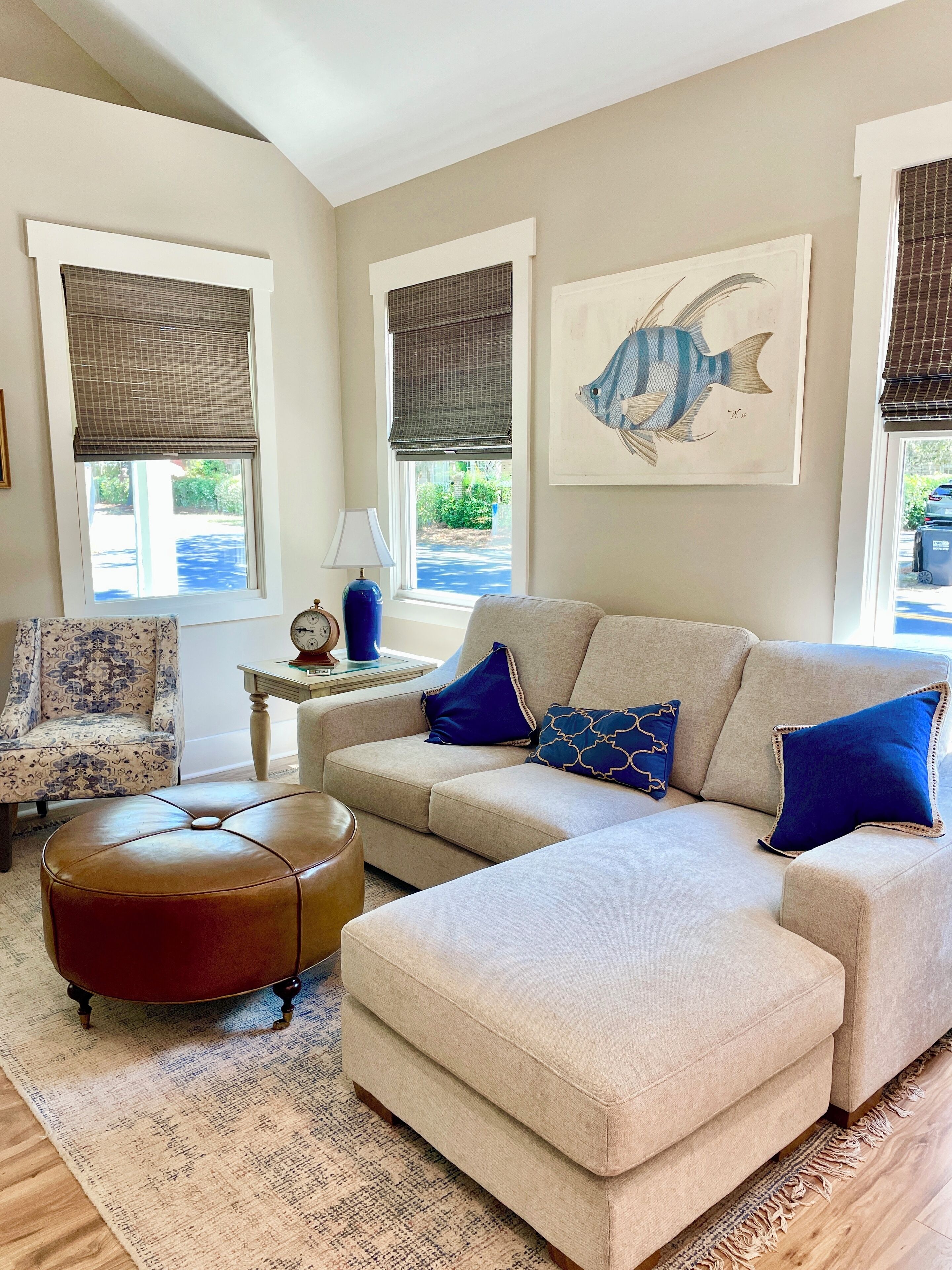 Una sala de estar con un sofá seccional color beige, un sillón estampado, un reposapiés redondo de cuero y una alfombra azul. Hay dos ventanas con persianas, un cuadro de peces colgado en la pared y una mesita con una lámpara.