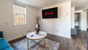 Een moderne woonkamer met een flatscreen-televisie aan de muur waarop “NETFLIX” wordt weergegeven, een ronde salontafel, een blauwe bank en een kamerkleed met een patroon.