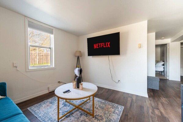 A modern living room with a flat-screen TV mounted on the wall displaying "NETFLIX," a round coffee table, a blue sofa, and a patterned rug.