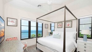 Een slaapkamer met een hemelbed, grote ramen met uitzicht op de oceaan, een nachtkastje en een stoel.