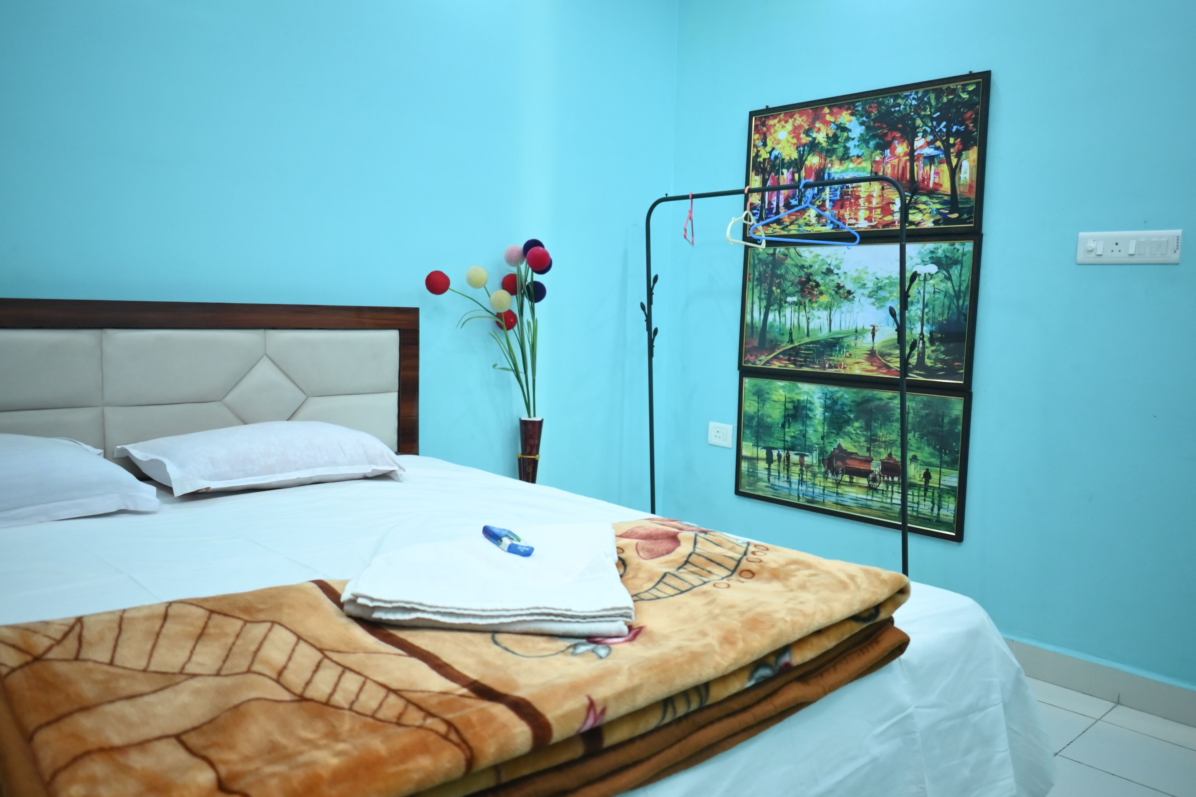 Ein ordentlich bezogenes Bett mit weißer Decke und brauner Wolldecke, eine Vase mit bunten Blumen sowie ein Gemälde einer Parkansicht an der Wand.