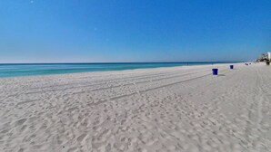 A wide sandy beach with footprints leading to the water, a clear blue sky, and a few buildings in the distance.
