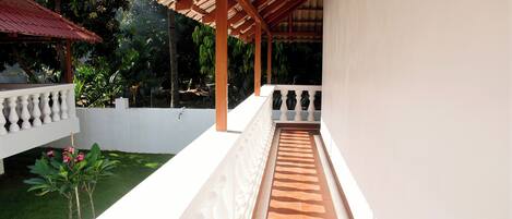 A covered balcony with a tiled floor, white balustrades, and a view of a garden with palm trees and a white building.