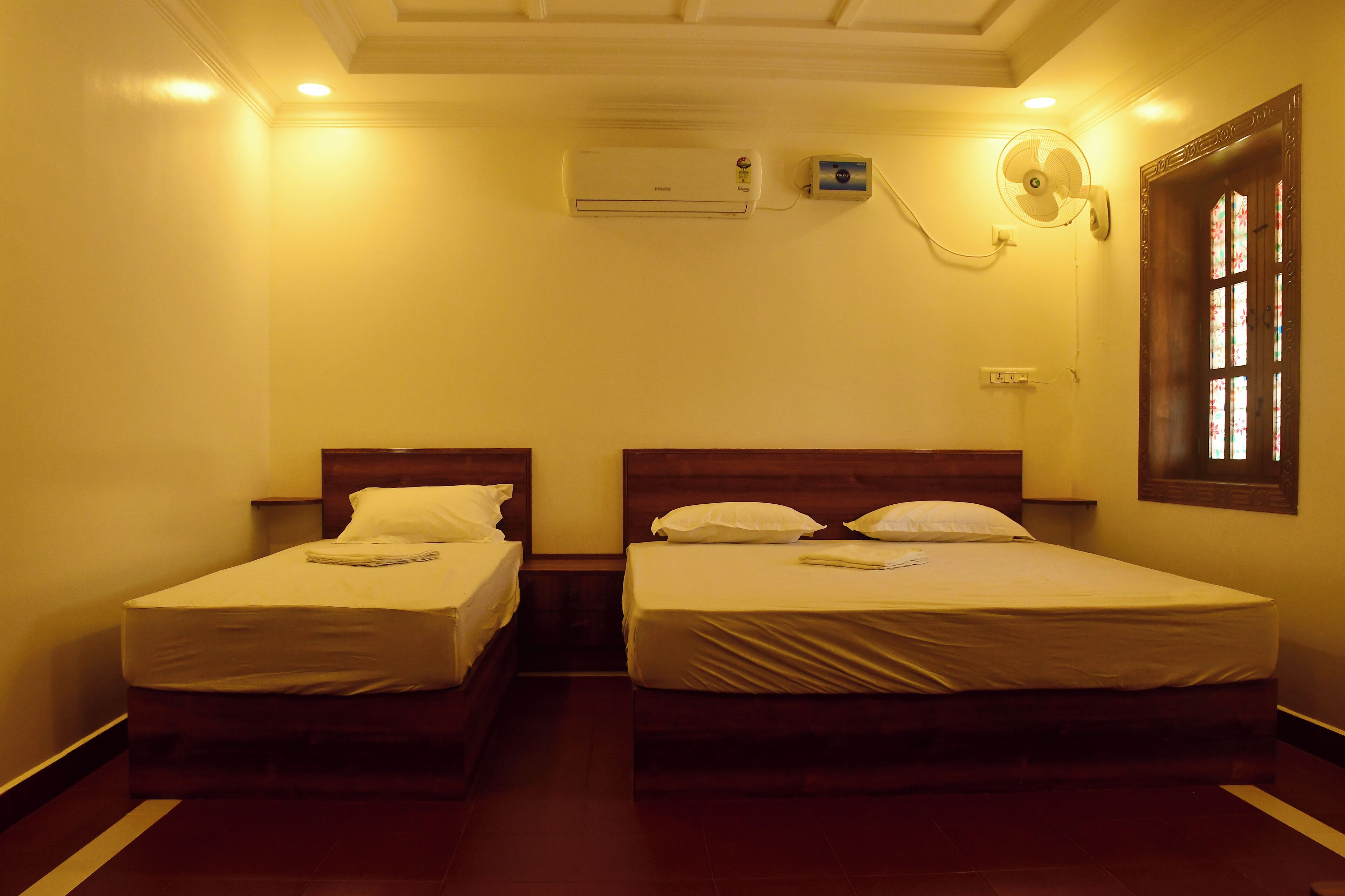 A hotel room with two single beds, a ceiling fan, an air conditioning unit, and a window with a decorative frame.
