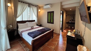 Een hotelkamer met een houten bed, een nachtkastje, een airconditioningapparaat, een kleine tafel met een lamp en een aan de muur bevestigde televisie.