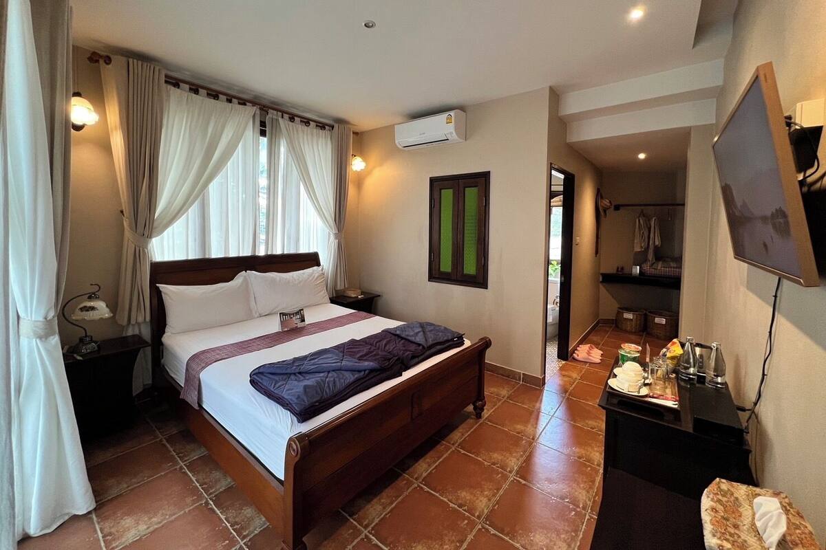 Camera d'albergo con un letto in legno, un comodino, un condizionatore d'aria, un piccolo tavolo con una lampada e una TV a parete.