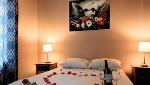 Ett hotellrum med en säng, två sänglampor, en tavla med motiv av fåglar och blommor på väggen och rosenblad utspridda på sängen.