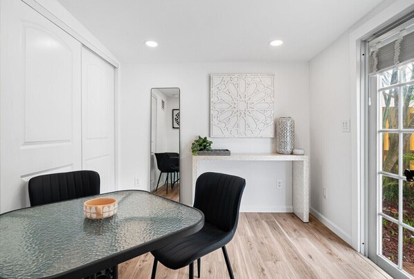A modern dining area with a glass table, black chairs, a white cabinet with decorative items, and a large window with a view of greenery.