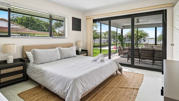 A modern bedroom with a large bed, a nightstand, a lamp, and a view of an outdoor patio with furniture. - Private pool home near downtown & beach (West Palm Beach)