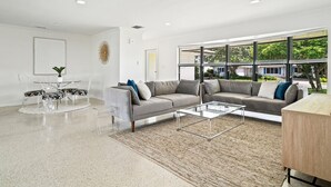 A modern living room with a grey sofa, a glass coffee table, and a dining area with a glass table and chairs. - Private pool home near downtown & beach (West Palm Beach)