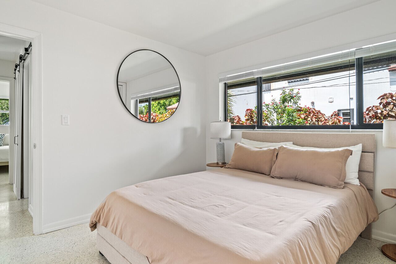Ein Schlafzimmer mit einem Bett, einem runden Spiegel, einer Nachttischlampe und einem Fenster mit Blick auf Bäume.