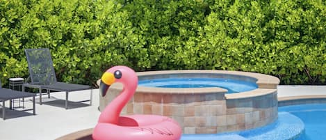 A swimming pool with a pink flamingo float and a hot tub, surrounded by green foliage.