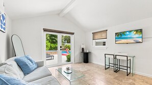 Una sala moderna con una mesa de centro de vidrio, un sofá con cojines azules, un televisor en la pared y vista a una piscina con un flotador de flamenco.