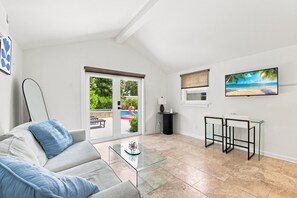 En moderne stue med et glasbord, en sofa med blå puder, et fjernsyn på væggen og udsigt til en swimmingpool med en flamingo-båd.