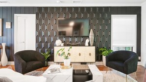 A modern living room with a dark patterned wall, a flat-screen TV, two dark armchairs, a white coffee table, and a wooden cabinet with decorative items. - Pool Home w/ Game Room | Near Peanut Island (West Palm Beach)
