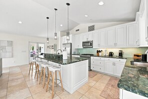 Una cocina moderna con armarios blancos, una isla central con taburetes, electrodomésticos de acero inoxidable e iluminación colgante.