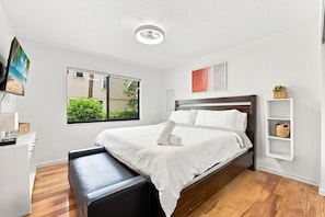 Habitación de hotel con cama, un banco, televisor, ventana con vista a la vegetación y una lámpara en el techo.