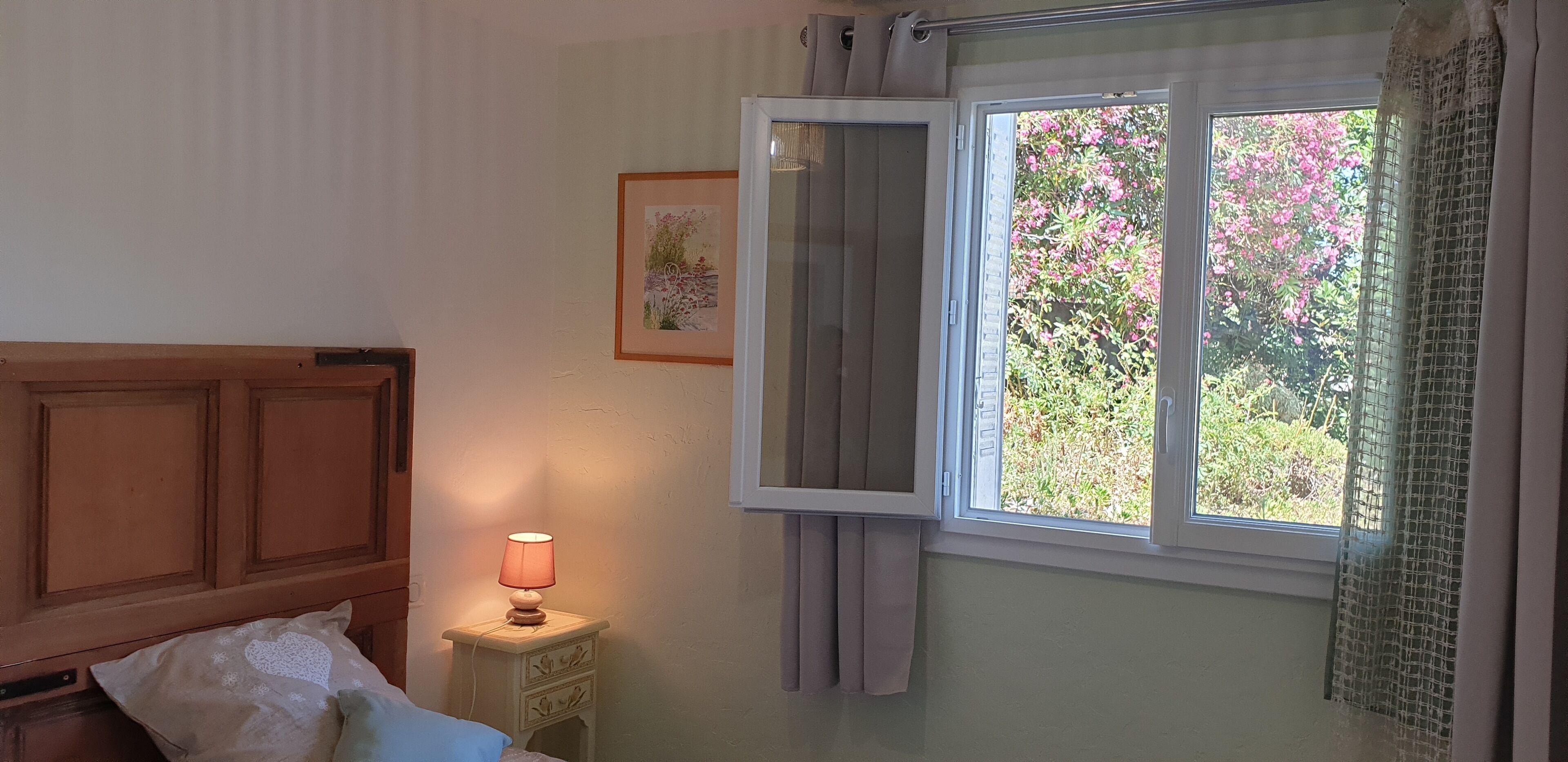 A bedroom with a wooden headboard, a bedside table with a lamp, a framed picture on the wall, and a window with a view of pink flowers.