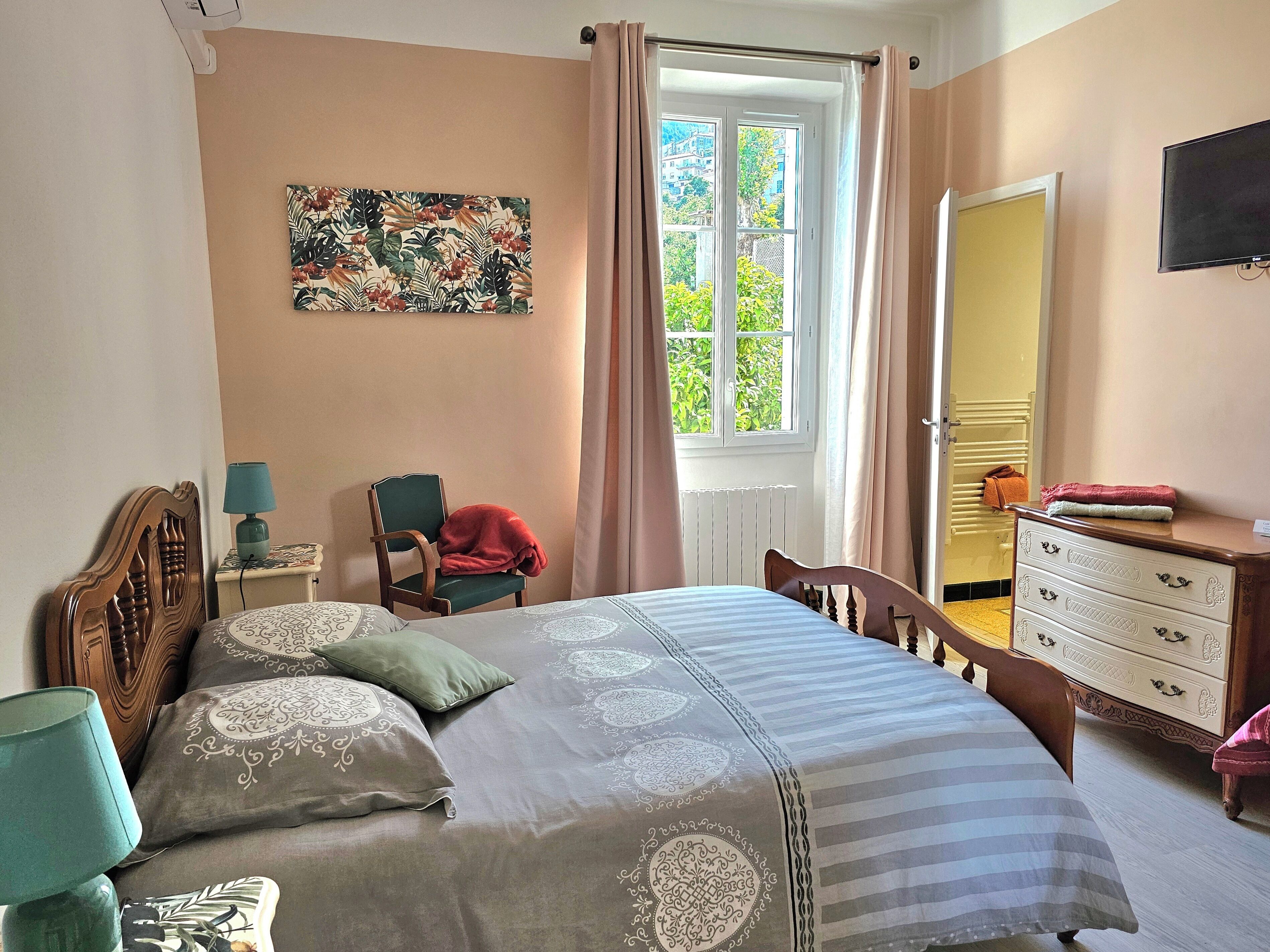 A bedroom with a bed, a chair, a dresser, a window with curtains, and a painting on the wall.