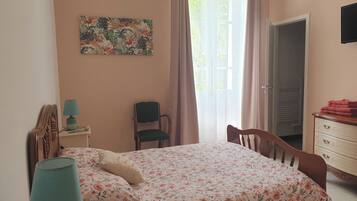 Un dormitorio con cama, una silla, una mesita de noche, una lámpara, un aparador, un cuadro y una ventana con cortinas.