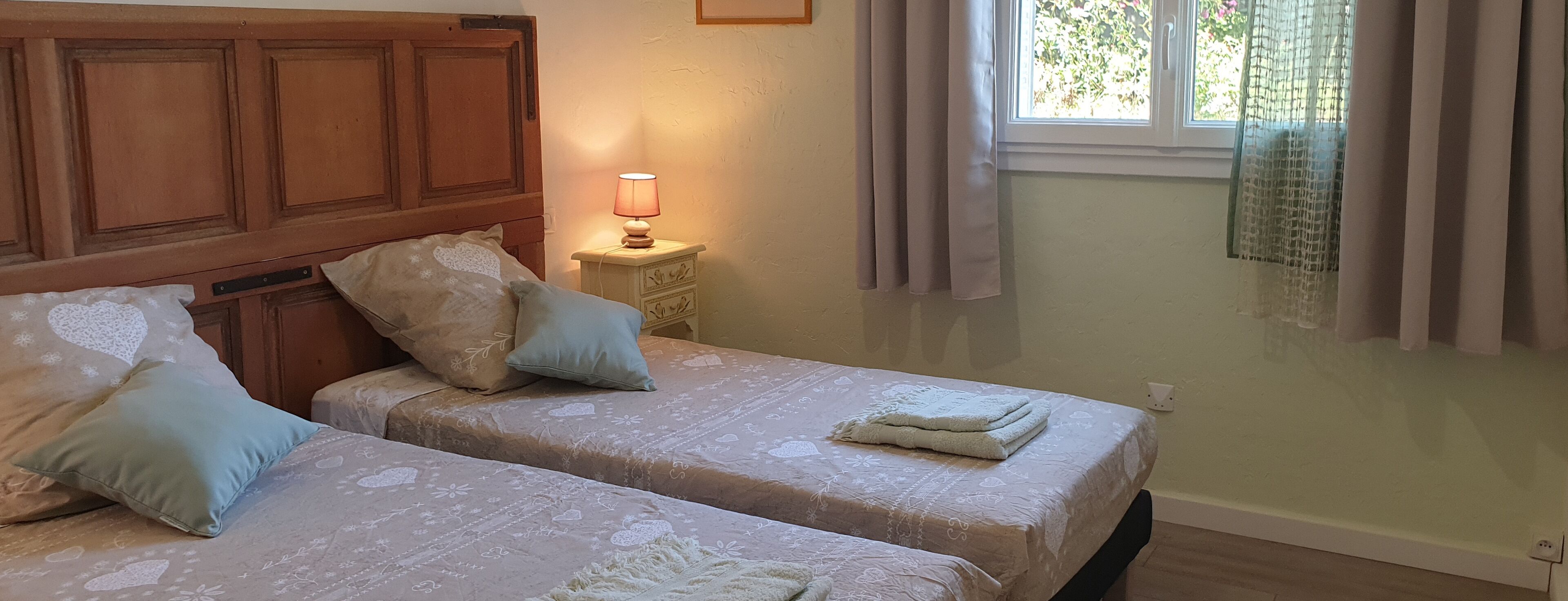 A room with two beds, a nightstand with a lamp, and a window with curtains.