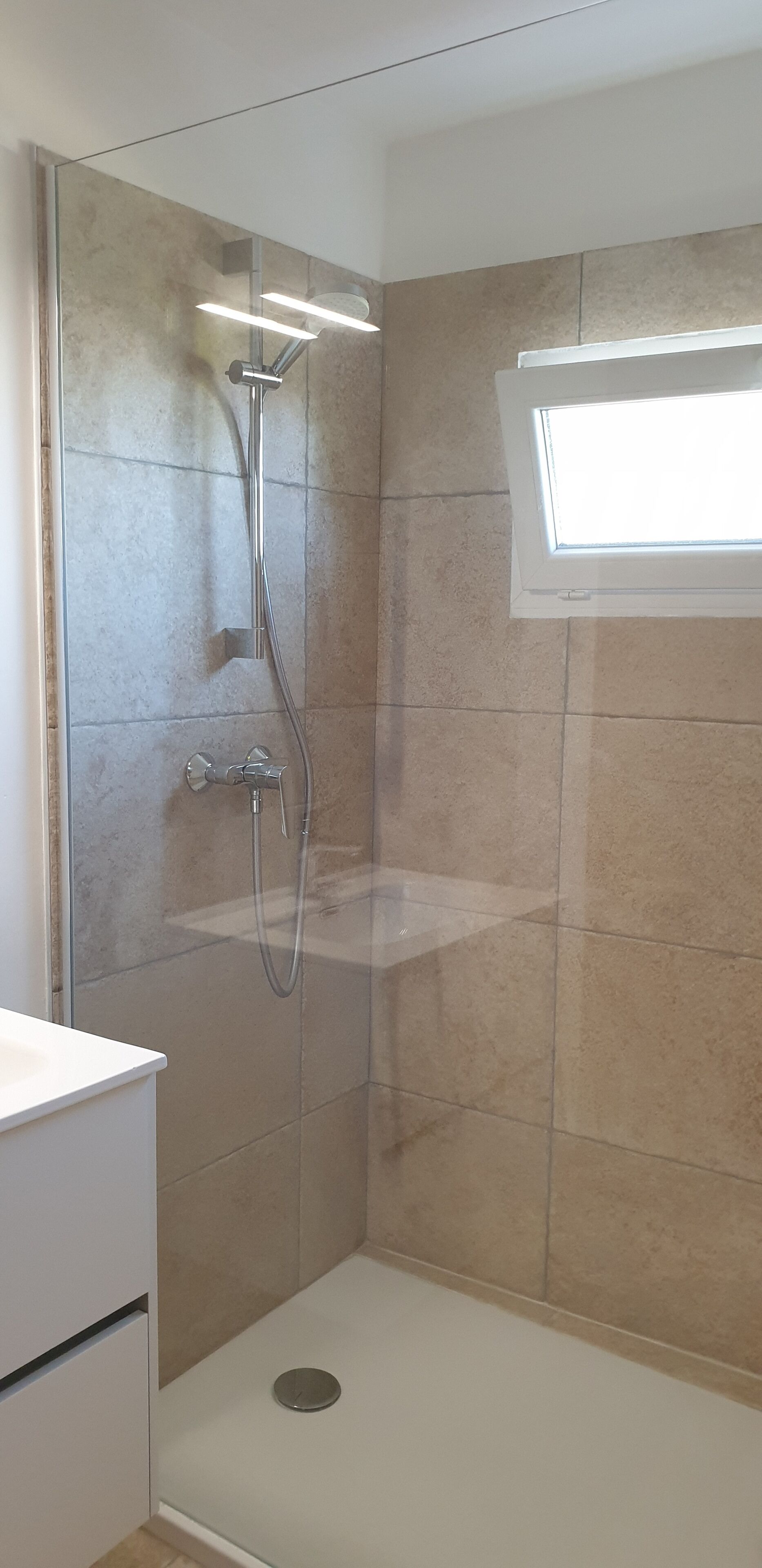 Una ducha con puerta de cristal, una ventana arriba y una encimera blanca a la izquierda.