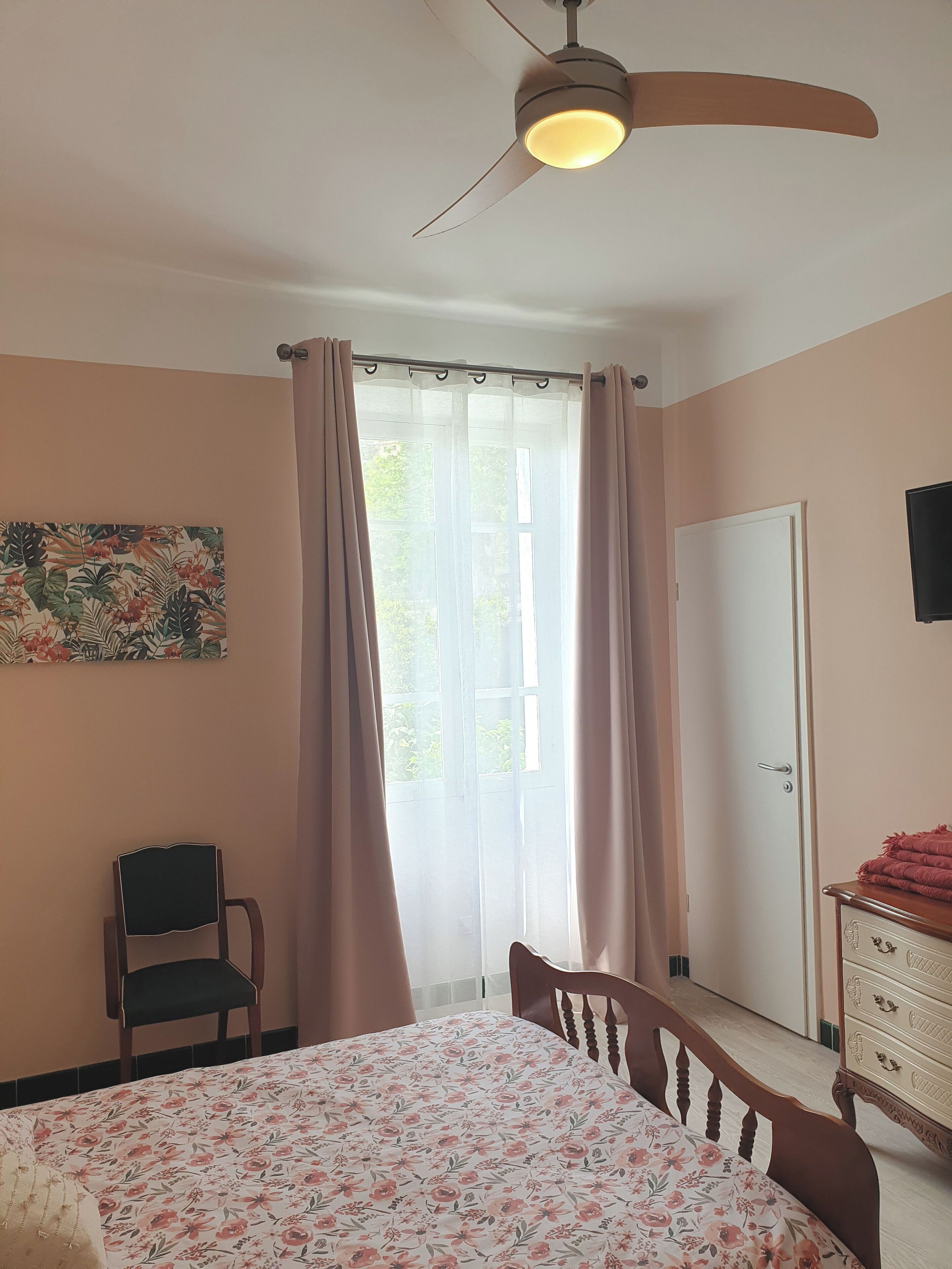 A bedroom with a bed, a chair, a dresser, a ceiling fan, a window with curtains, and a door.