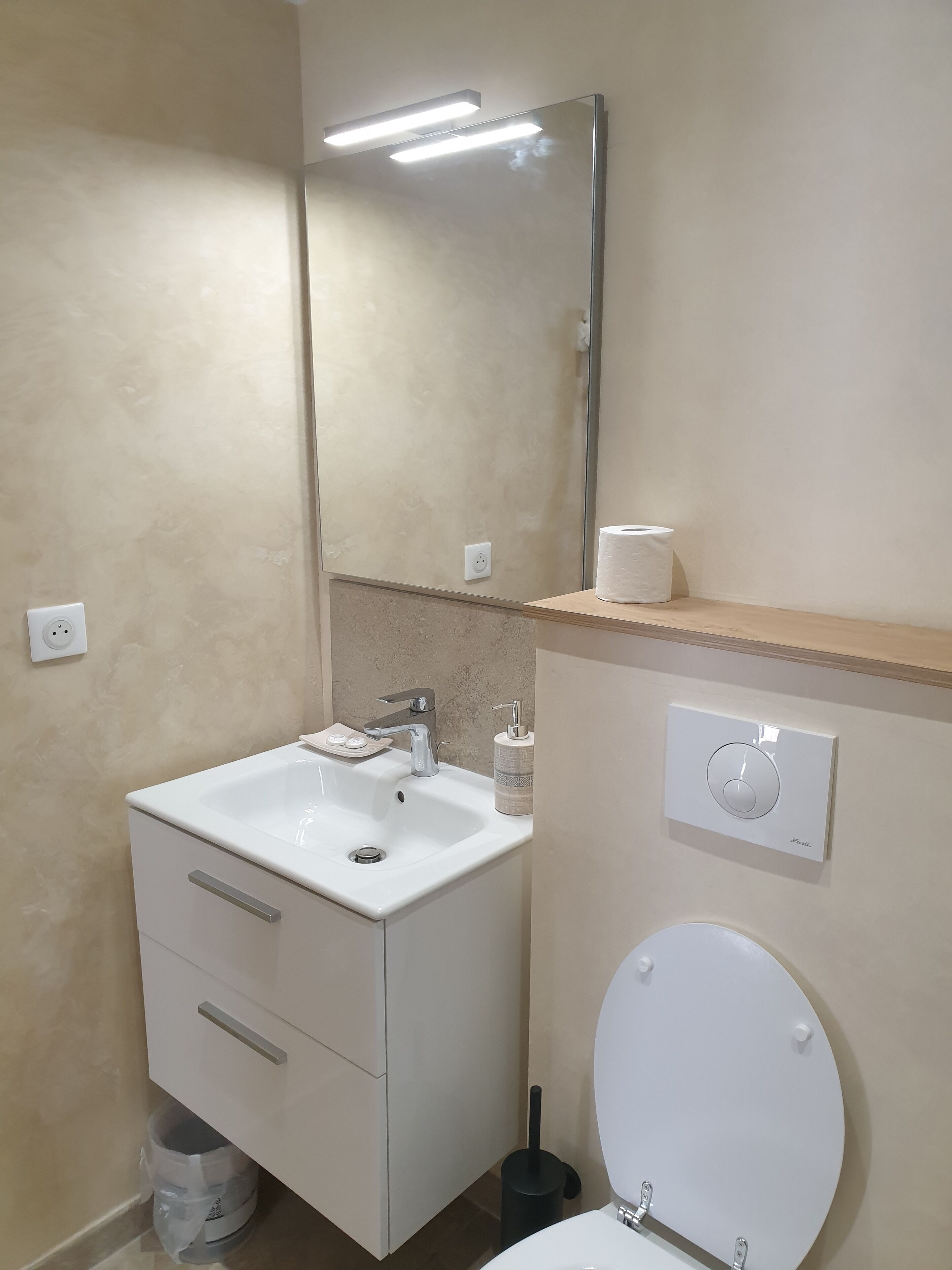 Une salle de bain avec un lavabo blanc, un miroir, des toilettes et une étagère sur laquelle se trouve un rouleau de papier toilette.