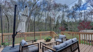 A wooden deck with outdoor furniture, including sofas and a table, surrounded by trees and a view of a lake.