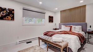 A modern bedroom with a large bed, a bench, a window with blinds, and artwork on the walls.