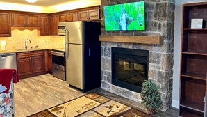 A modern kitchen with wooden cabinets, a stainless steel refrigerator, a stone fireplace, and a flat-screen TV mounted above it. - Cozy Riverside Studio in the Heart of Townsend (Townsend)