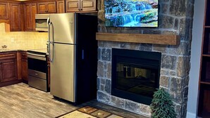 A modern kitchen with wooden cabinets, stainless steel appliances, a stone fireplace, and a flat-screen TV mounted above it. - Cozy Riverside Studio in the Heart of Townsend (Townsend)